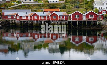 Cabanes de pêche traditionnelles en bois rouge (rorbu) sur des pilotis en bois se reflétant dans les eaux calmes du fjord d'un village de pêche historique, Lofoten, Norvège. Banque D'Images
