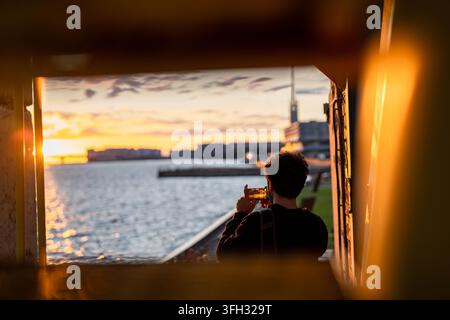 Homme non identifiable filmant une scène de coucher de soleil sur un pont et une baie de mer avec un smartphone, debout sur la côte. Banque D'Images