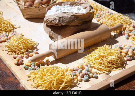 Ingrédients pour des pâtes maison avec un sac de farine, un rouleau à pâtisserie en bois, un panier à œufs et des nids de nouilles séchées disposés sur une planche rustique dans une cuisine naturelle et lumineuse Banque D'Images