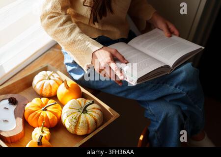 Adolescente lisant un livre près de la fenêtre avec des citrouilles à la lumière d'automne Banque D'Images
