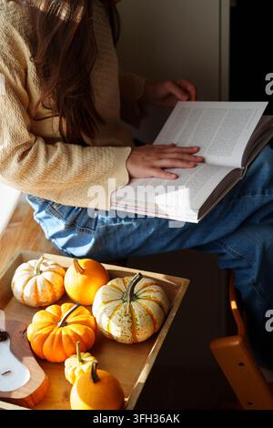 Adolescente lisant un livre près de la fenêtre avec des citrouilles à la lumière d'automne Banque D'Images
