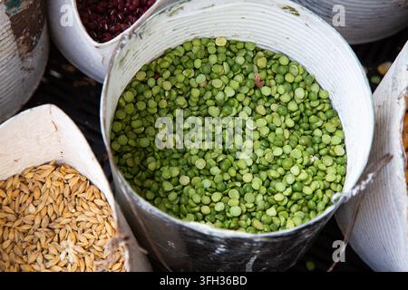 Légumineuses et grains séchés variés dans des contenants métalliques avec des pois cassés verts au centre, des haricots rouges et du blé visibles autour, texture rustique de nourriture de marché Banque D'Images