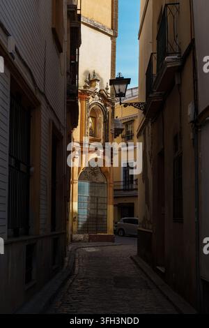 La lumière chaude du soleil illumine un shrine baroque orné intégré dans le coin d'un bâtiment jaune le long d'une ruelle pavée étroite, avec une lanterne suspendue a Banque D'Images