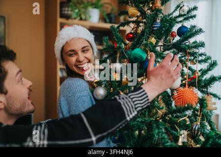 Couple décorant un sapin de Noël ensemble avec une femme portant un chapeau du Père Noël riant et souriant Banque D'Images