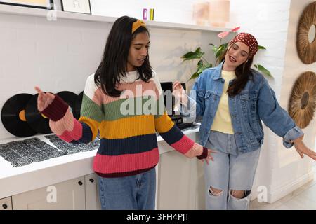 Différentes amies femmes dansant et bavardant à la maison près d'une platine avec des disques, portant des tricots à rayures Banque D'Images