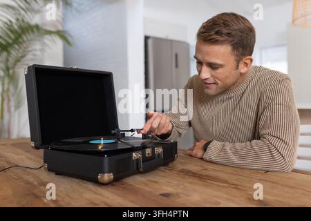 Homme adulte penché guidant le bras tonal vers un vinyle à étiquette bleue tournant sur une platine à valise dans la cuisine Banque D'Images
