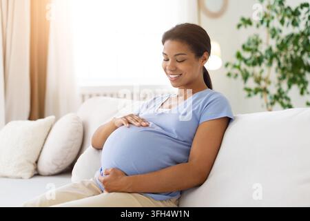 Femme noire enceinte se relaxant sur un canapé à la maison, touchant doucement son ventre Banque D'Images