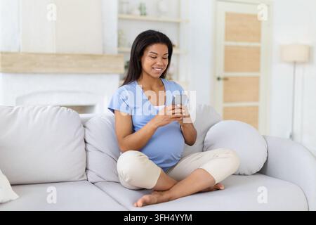 Femme noire enceinte souriante assise confortablement sur un canapé à la maison, utilisant un smartphone Banque D'Images