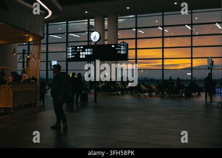 Passagers attendant dans un hall d'embarquement d'aéroport au coucher du soleil, silhouettés contre de grandes fenêtres panoramiques et un ciel orange brillant. Banque D'Images