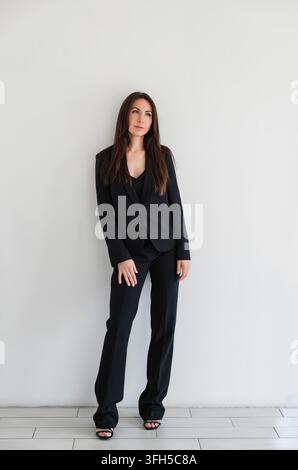 Femme regardant côté en pantalon ou costume formel noir se tient contre le mur blanc dans le studio. Modèle brune caucasien avec les cheveux longs. Portrait professionnel Banque D'Images