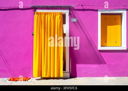Maison colorée rose et jaune avec un rideau jaune vif à Burano, lagune de Venise, Italie, avec des tomates séchant au soleil à l'extérieur. Banque D'Images