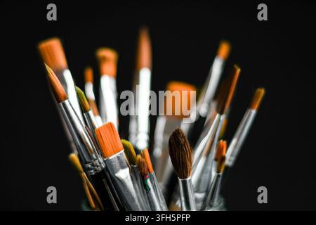 Assortiment de pinceaux d'artiste de formes et de tailles différentes disposés dans un récipient sur un fond noir foncé. Banque D'Images