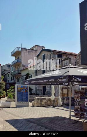 Pirovac, Croatie, 16 août 2026 Place méditerranéenne tranquille avec des maisons en pierre érodées, des balcons, des parasols de café et une vitrine de souvenirs sous un ciel éclatant Banque D'Images