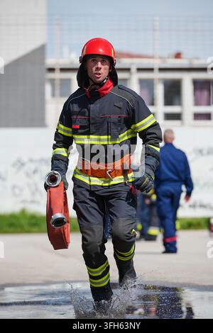 Sapeur-pompier courant avec un tuyau à travers des éclaboussures d'eau, portant un casque et un équipement de protection lors d'un exercice d'urgence Banque D'Images