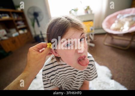 Un enfant espiègle tire la langue tandis qu'un adulte place une petite fleur jaune derrière son oreille dans un intérieur confortable, capturé d'en haut avec une prise de vue spontanée Banque D'Images