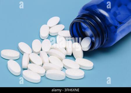 Comprimés blancs s'échappant d'une bouteille de médecine bleue posée sur une surface. Arrière-plan pharmaceutique pour le traitement des médicaments, les soins de santé, la pharmacie et la médecine. Banque D'Images