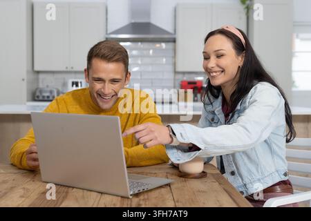 Couple assis dans la cuisine utilisant un ordinateur portable argenté, homme en pull jaune, femme avec bandeau rose pointe Banque D'Images