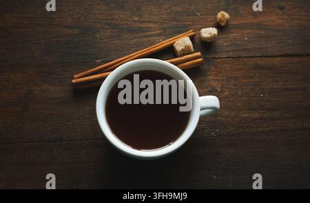 tasse en porcelaine blanche de thé ou de café, vue de dessus, avec bâtons de cannelle et morceaux de sucre brun sur bois Banque D'Images