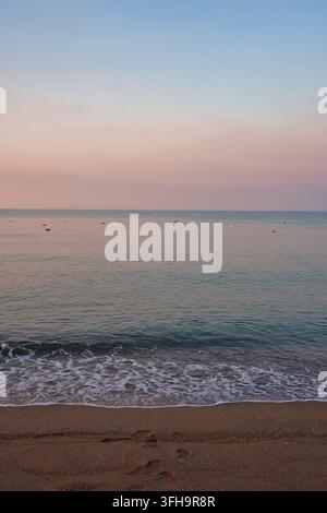 Un plan vertical d'une plage de sable sereine avec de douces vagues d'écume léchant la plage sous un ciel pastel rose doux au lever du soleil. De petites bouées flottent sur la Tra… Banque D'Images