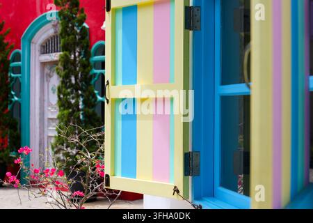 Une fenêtre ouverte colorée avec des volets pastel à côté d'une porte voûtée turquoise et d'un jardin fleuri, créant une façade résidentielle joyeuse avec des couleurs vives Banque D'Images