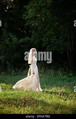 Une femme portant une longue robe blanche marche lentement à travers une zone herbeuse dans une forêt. Le soleil brille à travers les arbres pendant qu'elle avance. C'est la fin de l'après-midi. Banque D'Images