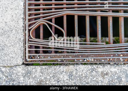 Vue rapprochée d'une grille métallique usée avec de la rouille, des barres linéaires et un détail décoratif courbé intégrée dans une surface en béton texturée. Banque D'Images