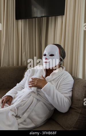 Femme afro-américaine allongée sur un canapé marron dans le salon, portant une robe de chambre blanche et un masque LED Banque D'Images
