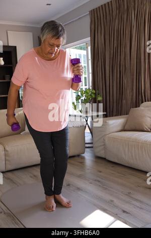 Femme afro-américaine âgée en tee-shirt rose et leggings tenant des haltères violets en équilibre à la maison Banque D'Images