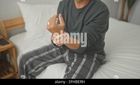 Homme d'âge moyen assis sur un lit tenant son poignet et grimçant dans une chambre à l'intérieur d'un bâtiment ; récupération de la douleur. Banque D'Images