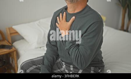 Homme serrant la poitrine avec sa main gauche sur un lit dans une chambre, assis droit, possible crise cardiaque ; inquiétude pour la santé. Banque D'Images