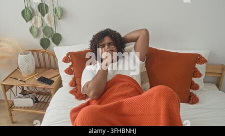 Homme allongé sur le lit avec une couverture orange serrant la poitrine et la main sur la gorge à côté de la table de chevet et smartphone dans le studio ; repos inconfortable. Banque D'Images