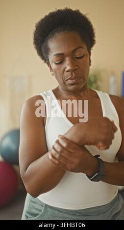 Femme massant son poignet et appuyant le pouce, montrant une douleur au poignet dans un studio de fitness à domicile portant un débardeur et une montre intelligente ; détermination. Banque D'Images