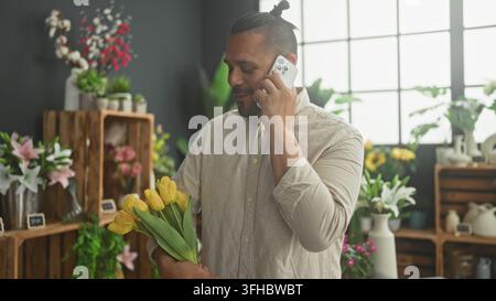 Un homme tient un bouquet de tulipes jaunes et parle au téléphone avec un smartphone tout en se tenant parmi des caisses en bois et des vases dans une fleuriste ; service calme et concentré. Banque D'Images