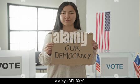 Femme tenant un panneau en carton lisant «sauvez notre démocratie» devant des isoloirs et une urne dans une salle de vote à l'intérieur d'un bâtiment ; détermination civique Banque D'Images