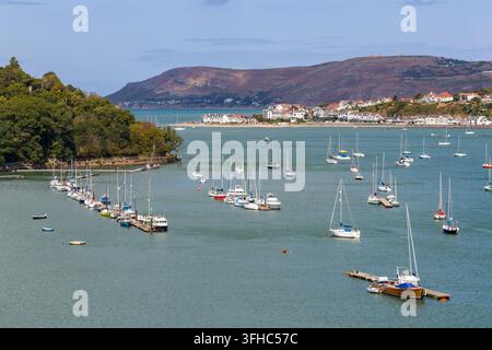 Vue en contre-plongée de nombreuses embarcations à voile ancrées sur une eau verte et calme, rive boisée à gauche et ville côtière lointaine sous des collines ondulées Banque D'Images