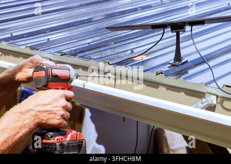 Le propriétaire utilise une perceuse pour fixer une gouttière près d'un panneau solaire sur un toit en métal Banque D'Images