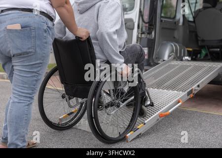 Une personne en fauteuil roulant reçoit de l'aide pour utiliser une rampe afin d'accéder à un van adapté. Un assistant personnel fournit un soutien dans une zone de stationnement. Banque D'Images