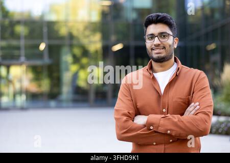 Succès professionnel et croissance de carrière, jeune homme confiant avec des lunettes debout devant un bâtiment de bureau moderne pour un portrait d'entreprise. Employé ambitieux posant les bras croisés. Banque D'Images