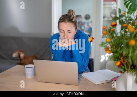 Freelance femme détendue et concentrée regardant l'écran de son ordinateur portable tout en travaillant depuis son bureau à domicile en télétravail. Banque D'Images