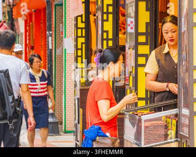 Macao, Chine, 16 avr. 2024 - Une femme en chemise rouge examine des cartons de viande séchée dans une vitrine en verre tout en interagissant avec un commerçant portant Banque D'Images