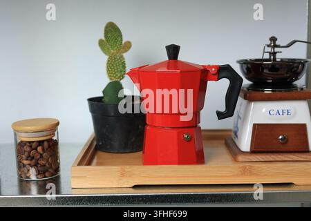 Moka expresso rouge sur un plateau en bois. Une plante cactus ajoute une touche verte à côté. Les grains de café sont stockés dans un pot transparent avec couvercle en bambou. Griß à manivelle Banque D'Images