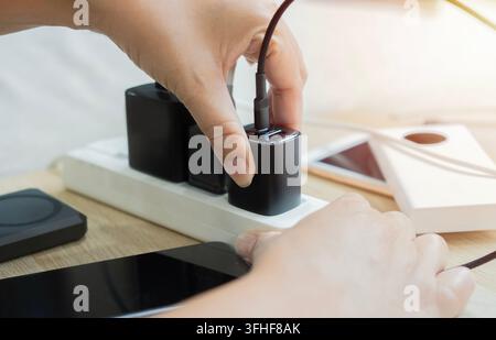 Gros plan en contre-plongé d'une personne branchant ou débranchant un bloc de chargeur de téléphone noir d'une multiprise sur un poste de travail propre. Banque D'Images