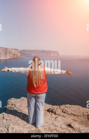 Femme en liberté debout sur une falaise rocheuse, les bras ouverts, surplombant la mer bleue et les montagnes lointaines avec espace libre Banque D'Images