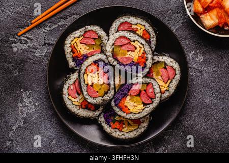 Une assiette noire remplie de kimbap (ou gimbap) aux garnitures colorées. Les ingrédients comprennent des saucisses, des cornichons, des œufs et des légumes. Banque D'Images