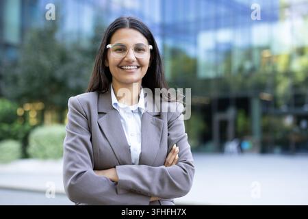 Femme professionnelle en costume d'affaires, leader féminine confiante avec des lunettes souriant à l'extérieur devant un bâtiment de bureaux d'entreprise. Succès professionnel et leadership. Banque D'Images