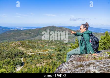 Un randonneur avec un sac à dos est assis sur un point de vue rocheux, pointant vers les collines verdoyantes sous un ciel bleu clair. Banque D'Images