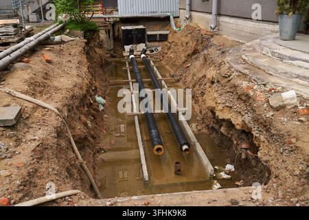 Tranchée de construction pour la rénovation urbaine dans une cour à côté d'un bâtiment avec deux gros tuyaux noirs enjambant une excavation boueuse sur des supports en bois et Banque D'Images