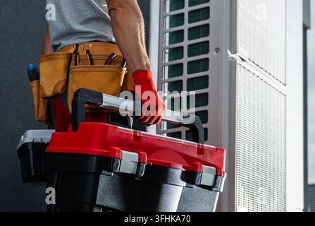 Un ouvrier porte des boîtes à outils à côté d'une unité extérieure de climatisation tout en portant des gants et une ceinture à outils. Banque D'Images