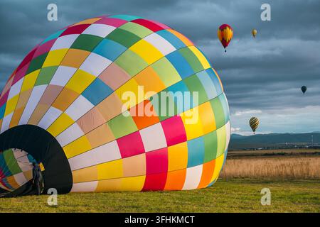 Enveloppe de ballon aerostatique coloré à carreaux posée sur un champ d'herbe verte pendant la préparation du gonflage avec des ballons flottants au loin sous un ciel nuageux Banque D'Images