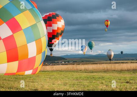 Grand ballon à air chaud coloré à carreaux au premier plan sur le champ avec un ballon rouge qui décolle et des ballons flottants contre un ciel nuageux sur une vaste montagne Banque D'Images